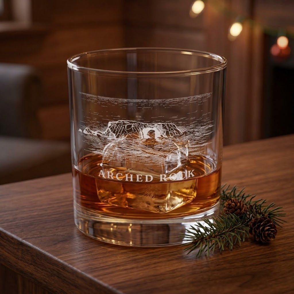 Arched Rock engraved rocks glass with whiskey and ice on a wood bar, holiday lights and an evergreen sprig behind