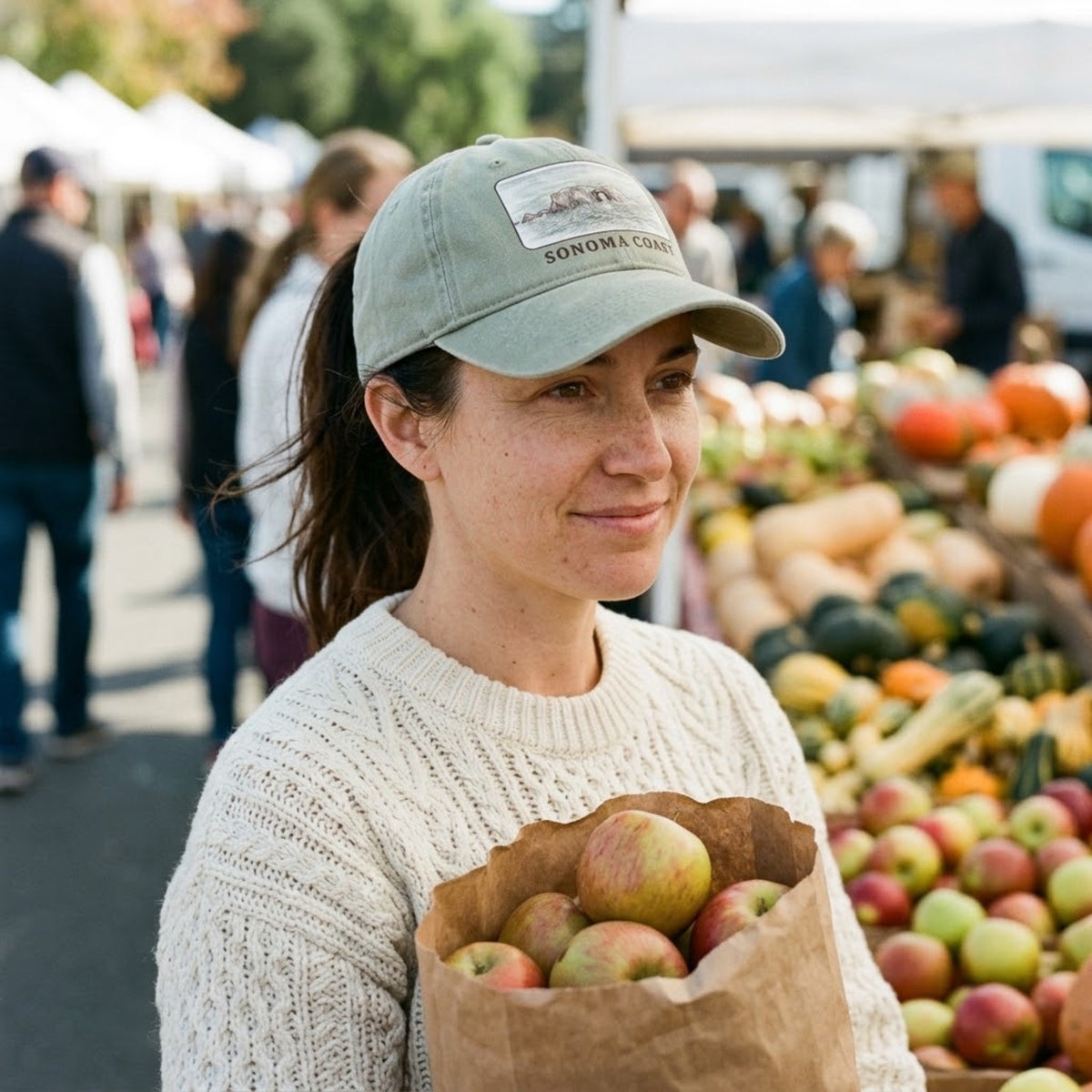 Woman wearing the Sandstone Arched Rock washed cap at a Sonoma County farmers market, holding a bag of apples